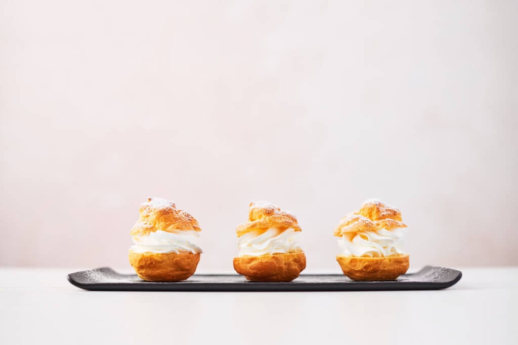 Delicious cream puff cakes with cream and powdered sugar on a black plate on white marble table over pink background.