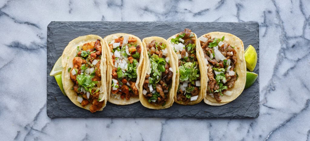 row of mexican street tacos on slate with carne asada and al pastor top down