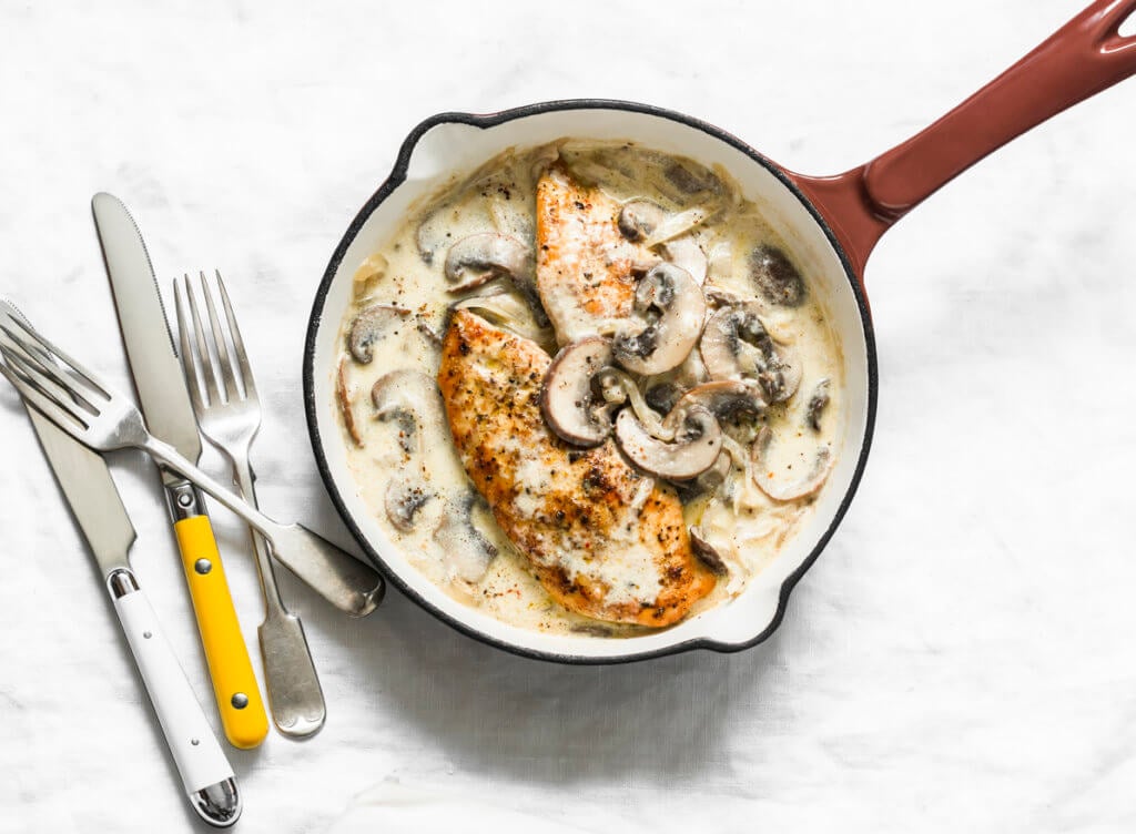 Roasted chicken fillet with cheesy mushrooms cream sauce in a cooking pan on a light background.