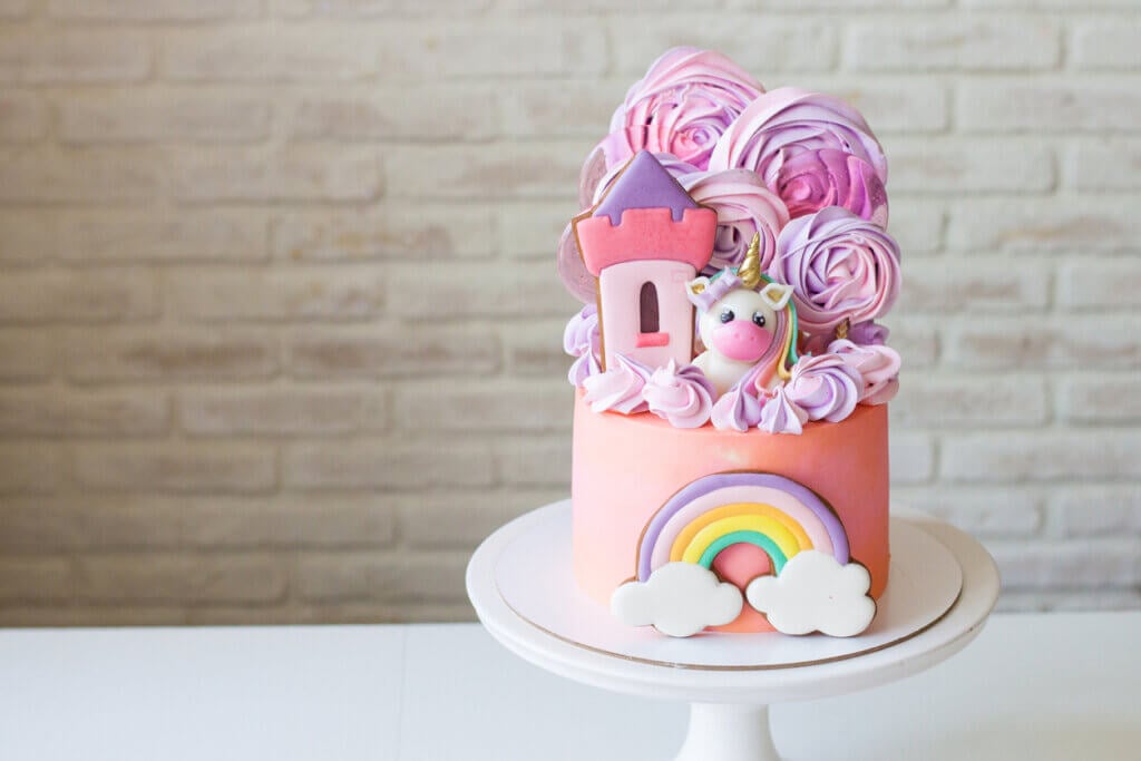 Cute pink birthday cake for a little girl with fondant unicorn, gingerbread princess castle, rainbow and meringue clouds.
