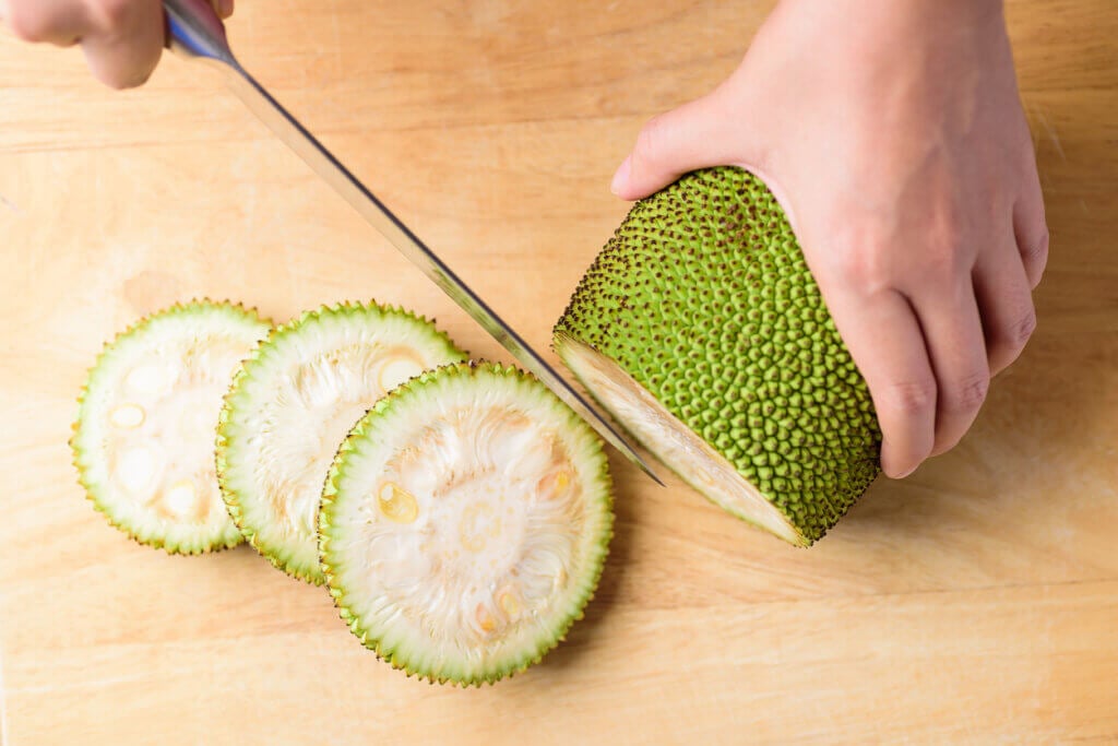 Hand holding kitchen knife and cutting young jackfruit on wooden board for cooking, 