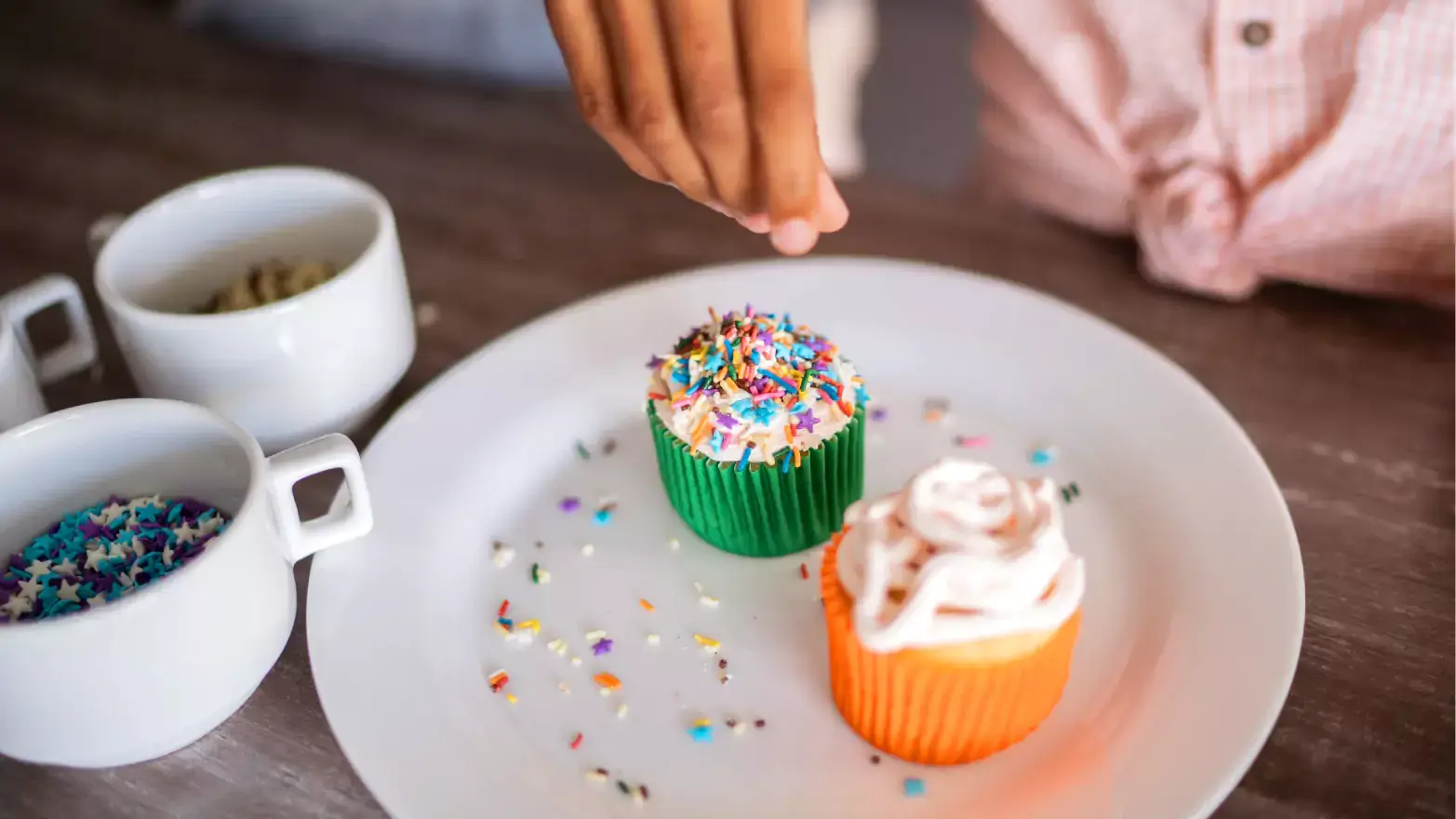 Child sprinkling rainbow sprinkles on cupcake with white frosting