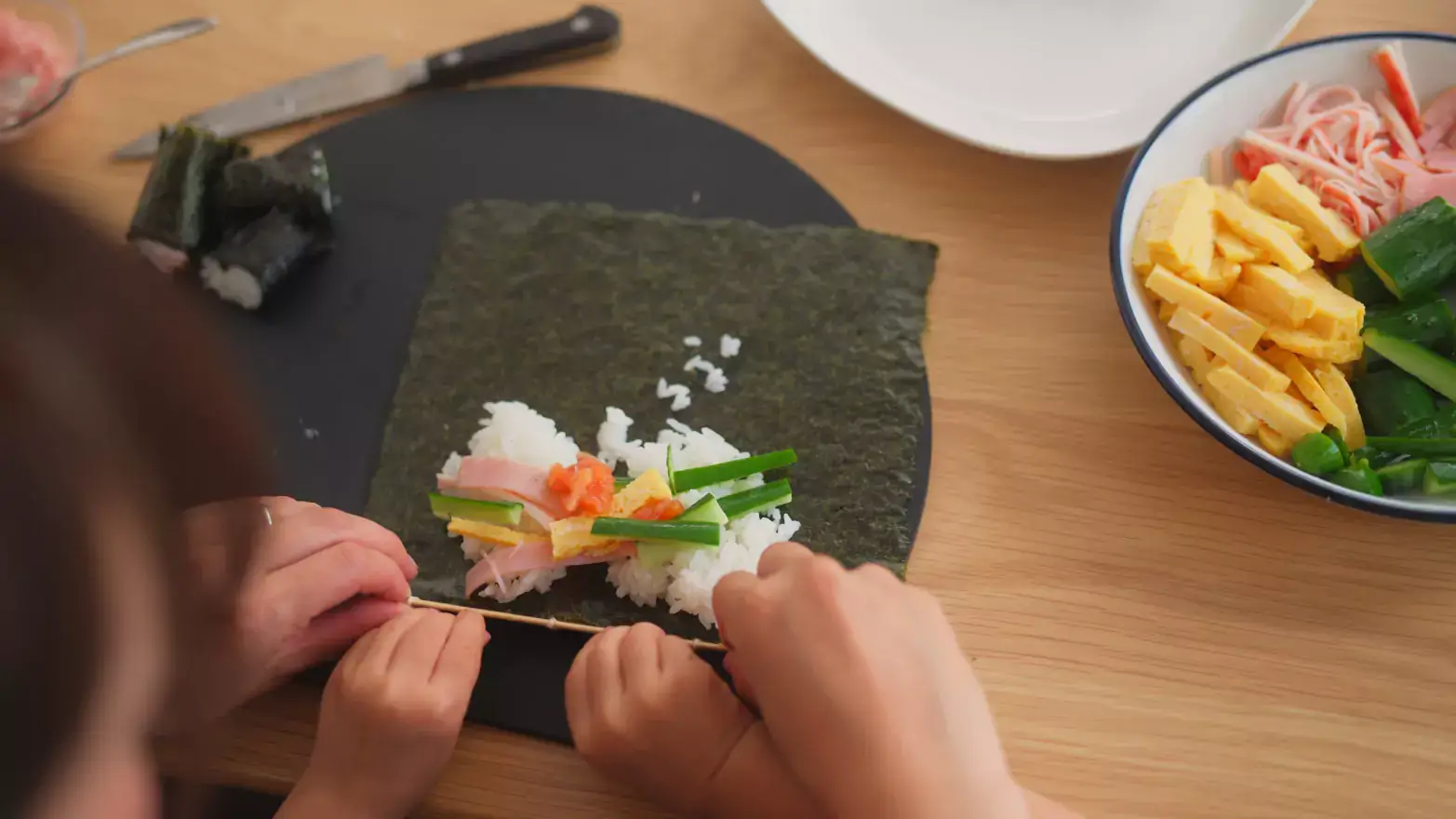 Parent helping child roll a sushi roll with rice, cucumber, tofu and spam